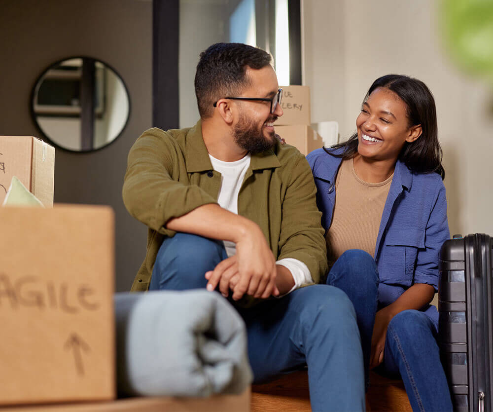 A smiling couple sitting among moving boxes and luggage, planning their next home purchase before selling their current one.