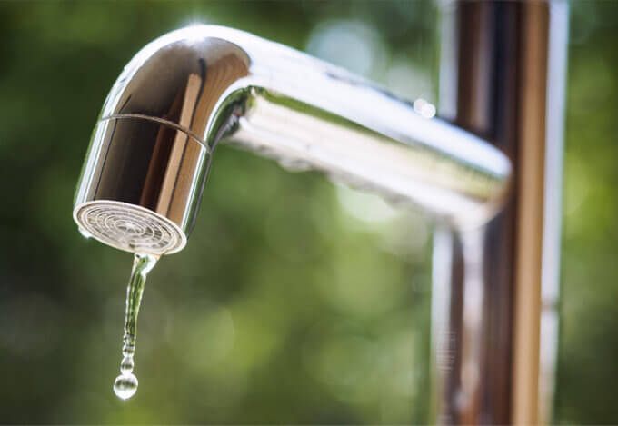 Close-up of a metal faucet with a water droplet hanging from the spout, symbolizing monthly utility expenses.