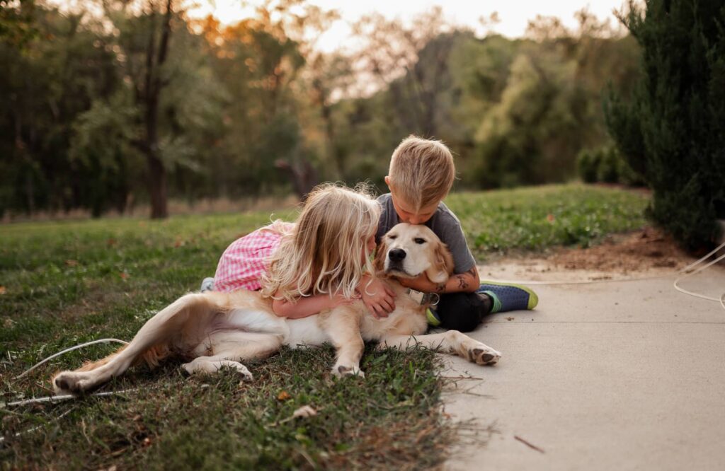 Two kids sitting outside their home hug their dog.