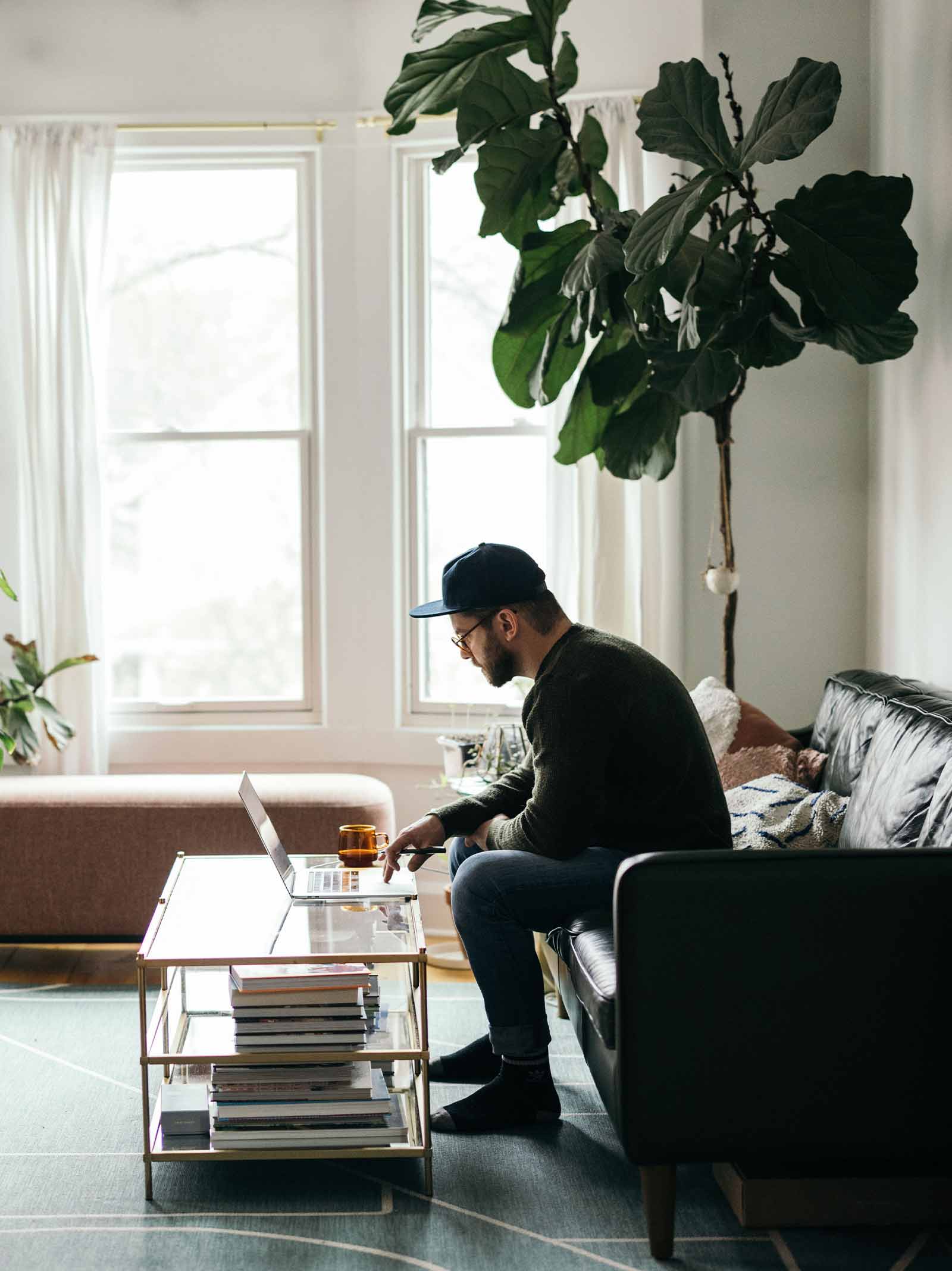 Man in living room sits on couch reaching refinancing guide on his laptop computer.