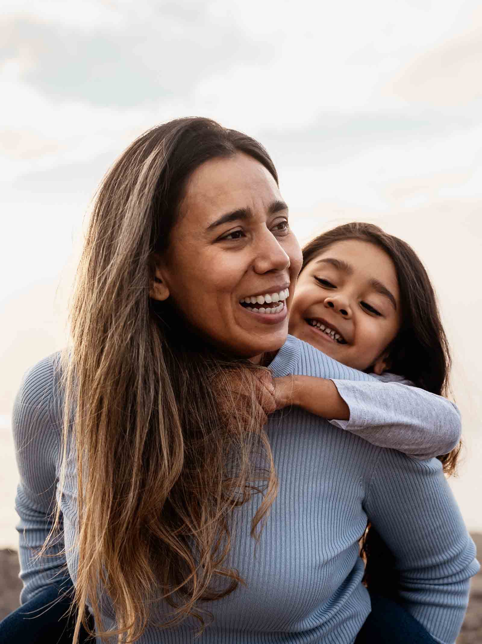 Mother with daughter on her shoulders smile together near the ocean.